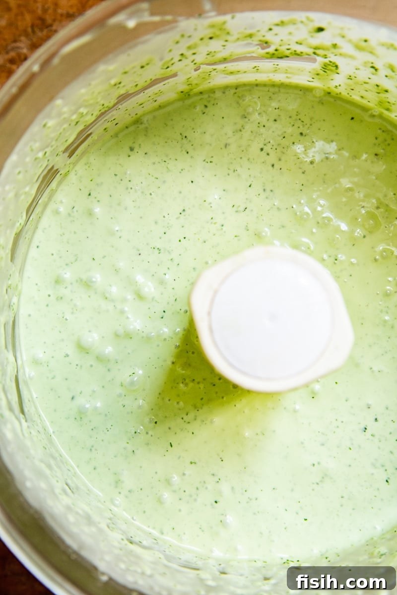 A close-up view of fresh green herbs, possibly basil and parsley, being blended in a food processor, destined for a vibrant Green Goddess dressing.