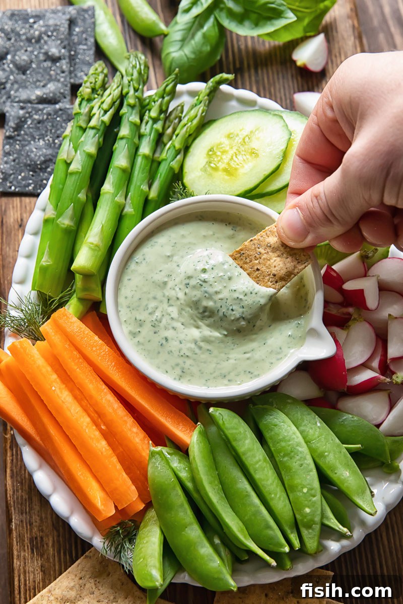 A cracker being dipped into a bowl of vibrant green goddess dip, with a platter of assorted fresh vegetables in the background.