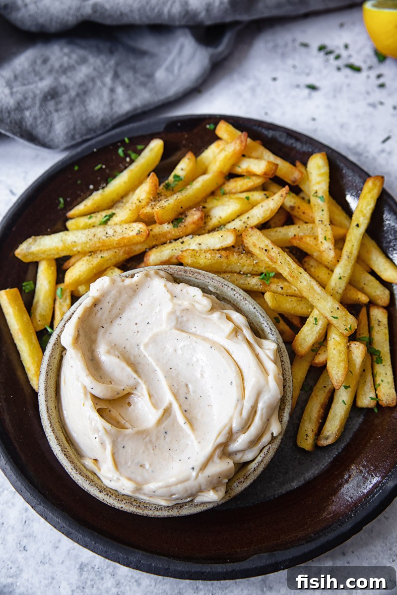 Top down view of truffle aioli in a white bowl, surrounded by golden, crispy fries on a rustic plate.