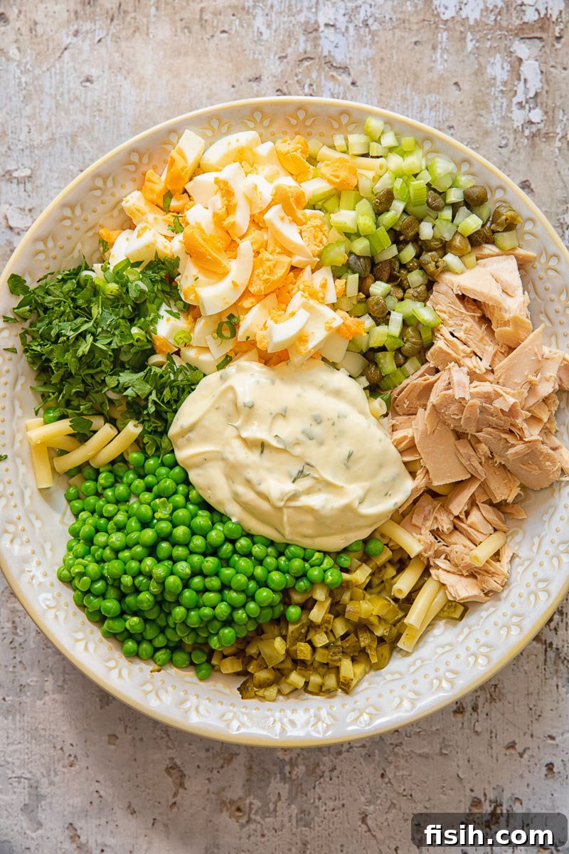 A process shot showing various fresh ingredients for tuna macaroni salad, including chopped celery, peas, and dill pickles, ready to be combined in a bowl.