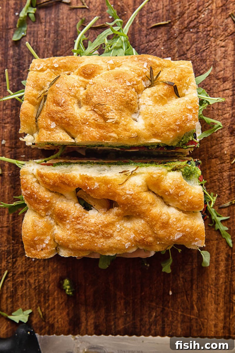 Top-down view of a large focaccia sandwich, expertly cut in half to reveal its vibrant layers of Italian deli meats, cheese, and vegetables on a wooden cutting board.