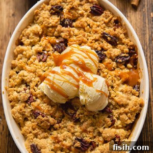 Top down view of a scoop of vanilla ice cream melting on a warm apple crisp, garnished with toasted pecans, in a ceramic baking dish.