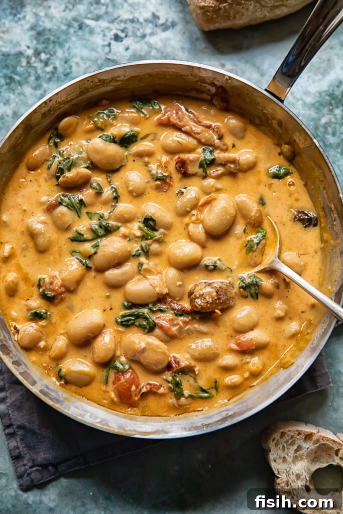 Top down shot of a spoon scooping tuscan butter beans in sun-dried tomato sauce with spinach out of a pan