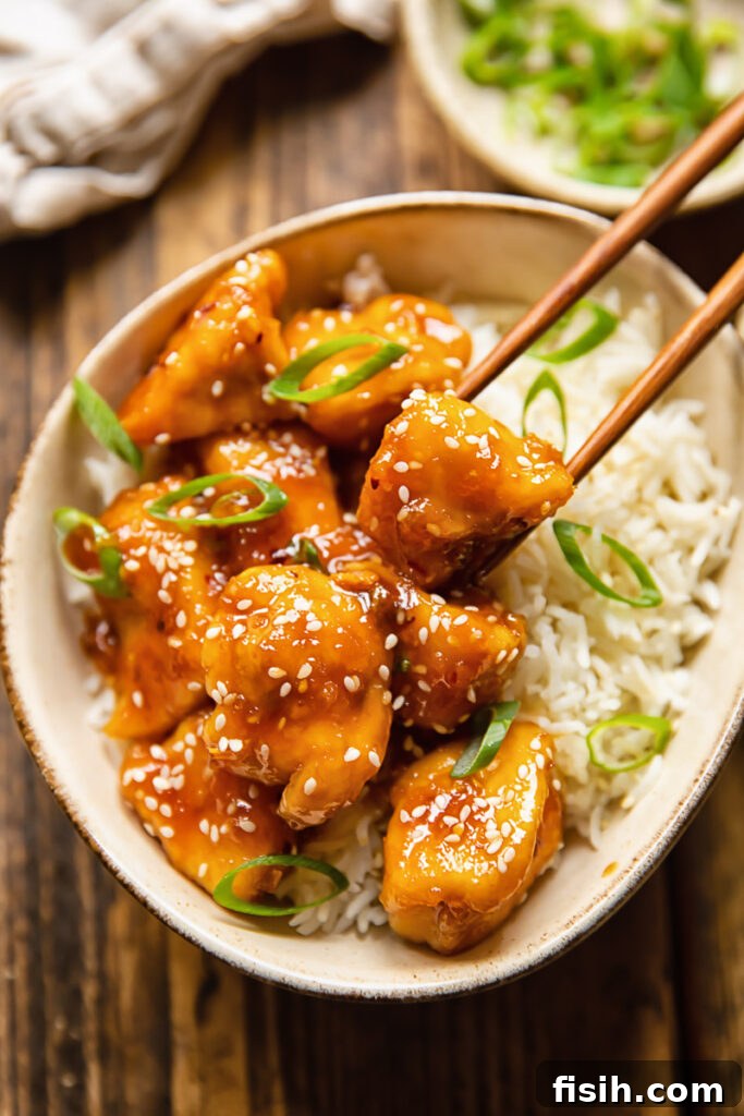Chopsticks removing a piece of honey sesame chicken from a bowl of rice, highlighting the glossy glaze and sesame seeds.
