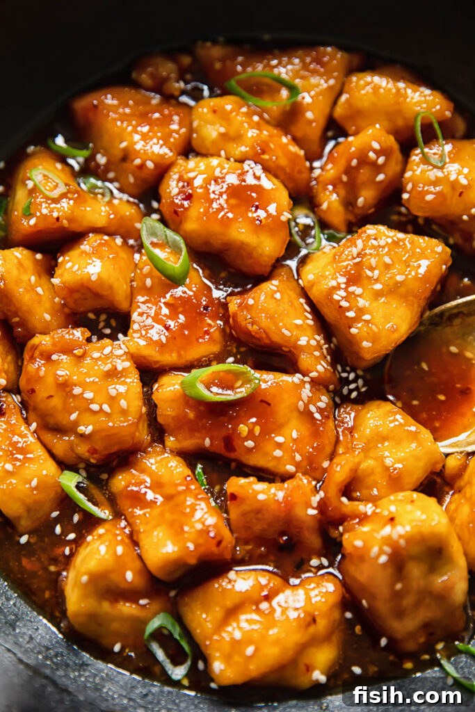 Close up view of glossy honey sesame chicken garnished with sesame seeds and green onions.