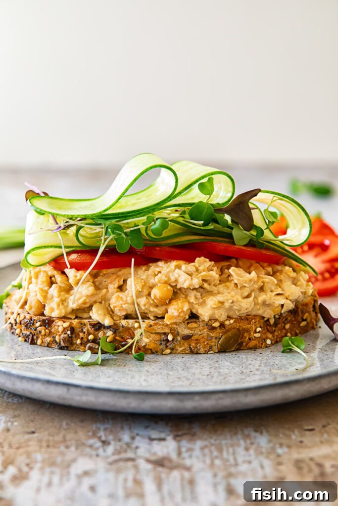 Smashed Chickpea Sandwich 3 A chickpea salad sandwich with its top slice open, revealing a generous layer of creamy chickpea filling and fresh vegetables on a plate.