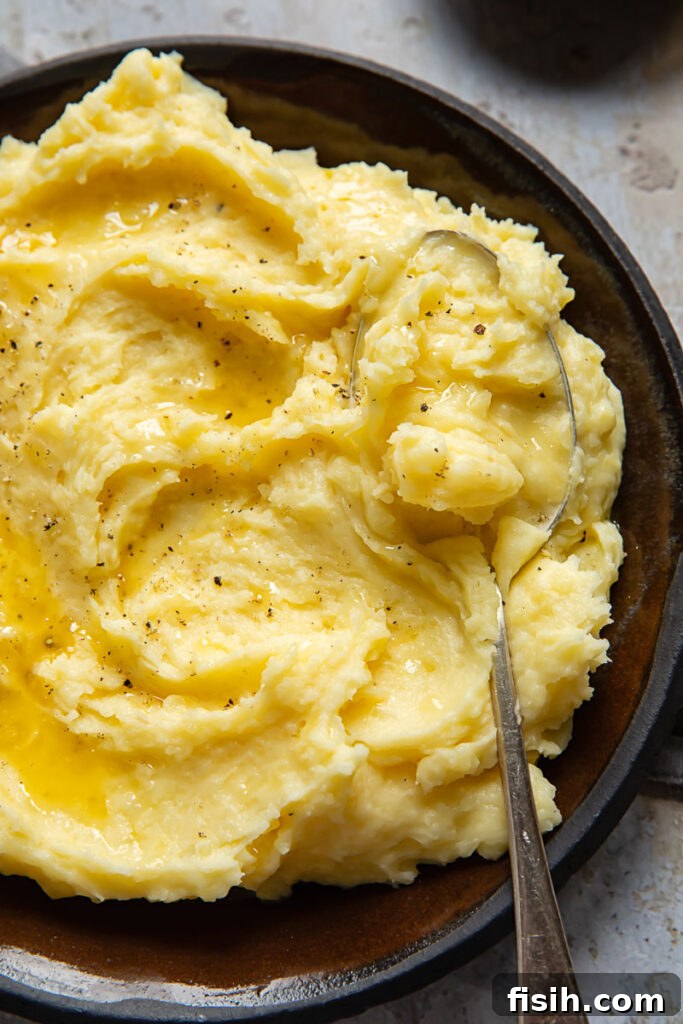 Close up shot of creamy Parmesan mashed potatoes on a white plate with a spoon, topped with a dollop of melted butter and a sprinkle of black pepper