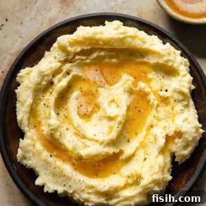 Top-down view of a generous serving of creamy, perfectly smooth Brown Butter Mashed Potatoes, beautifully garnished with a sprinkle of fresh green herbs. An elevated take on a classic side.