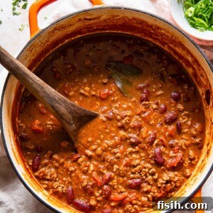 Top down view of a rich, earthy-red beef and kidney bean chili con carne in an orange pot, garnished with fresh cilantro and a dollop of sour cream.