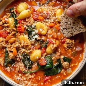A hearty bowl of Sausage Gnocchi Soup with 'Nduja, showing a hand dipping a piece of crusty bread into the rich broth, highlighting tender gnocchi and sausage pieces.
