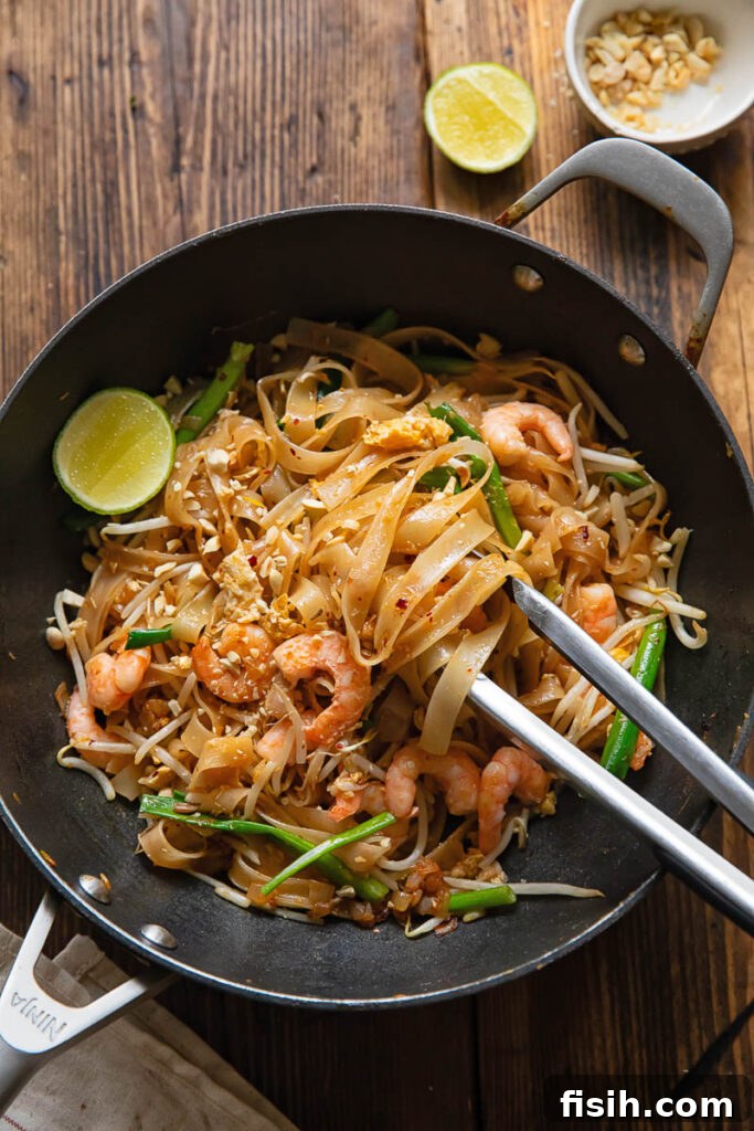 A steaming wok filled with delicious Shrimp Pad Thai, tongs removing a generous portion, alongside fresh lime wedges and crushed peanuts, ready to be served.